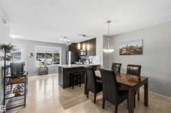 Open-concept living area with light wood-finish flooring, featuring a kitchen island with granite countertop and a dining space with a contemporary pendant light fixture - 