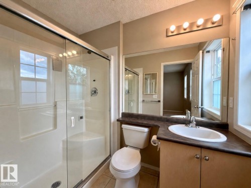 Bathroom featuring a glass-enclosed shower with integrated bench seating, a single-sink vanity with a dark countertop, a wall-mounted mirror, and a four-bulb vanity light fixture - 12207 16 Avenue, Edmonton, AB 