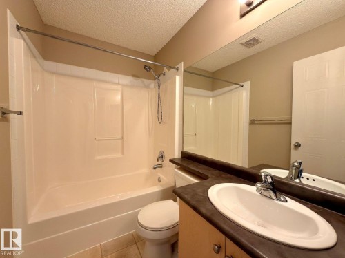 Bathroom featuring a shower-tub combination with a curved shower rod, a white toilet, and a vanity with an integrated sink, dark countertop, and base cabinetry - 12207 16 Avenue, Edmonton, AB 