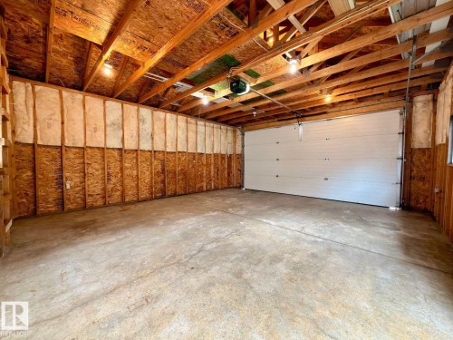 Spacious garage interior featuring exposed wood beams, concrete flooring, a large white sectional garage door, and visible insulation along the wall framing - 12207 16 Avenue, Edmonton, AB 