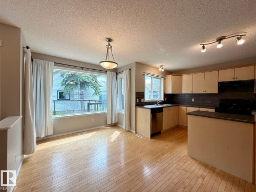 Open-concept kitchen and dining area featuring wood-finish flooring, a large picture window, a built-in kitchen island, stainless steel dishwasher, and track lighting - 12207 16 Avenue, Edmonton, AB 