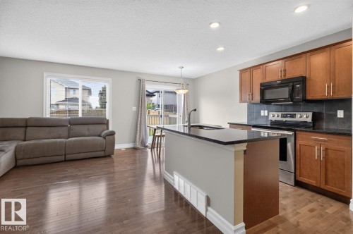 Open-concept living area with dark wood-finish flooring, a kitchen island with a dark countertop, stainless steel appliances, wood cabinetry, and a sliding glass door leading to an exterior deck - 12032 167B Avenue, Edmonton, AB 