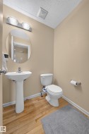 Pedestal sink and toilet with light wood-finish flooring, a round wall mirror, and a two-light vanity fixture - 