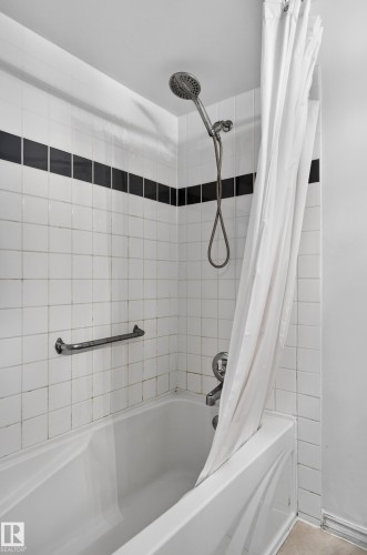 Bathtub and shower combination featuring white square tiling with a black accent band, a shower head with hose attachment, and a chrome grab bar - 13445 40 Street Nw, Edmonton, AB 