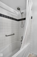 Bathtub and shower combination featuring white square tiling with a black accent band, a shower head with hose attachment, and a chrome grab bar - 