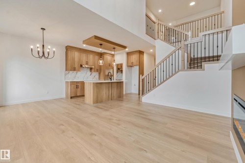 Open-concept living space featuring wood-finish flooring, a contemporary kitchen with light wood cabinetry and a central island, a two-story ceiling, and a prominent staircase with iron balusters - 4636 18 Avenue, Edmonton, AB 