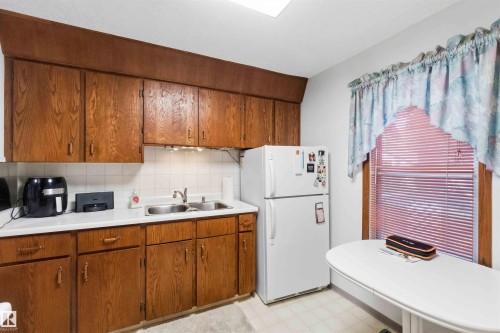 Kitchen featuring wood cabinetry, white countertops, a double basin stainless steel sink, and a white tile backsplash - 12016 61 Street, Edmonton, AB 