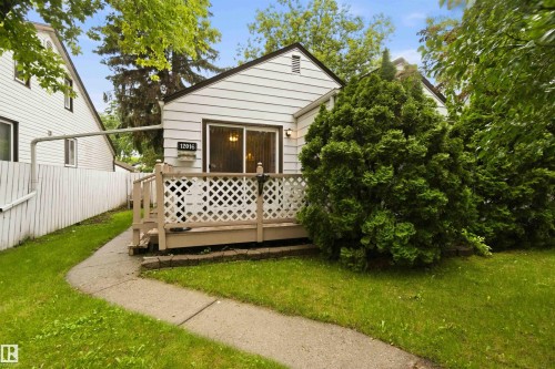 White siding exterior featuring a front deck with lattice railing, mature shrubs, a concrete pathway, and a picket fence - 12016 61 Street, Edmonton, AB 