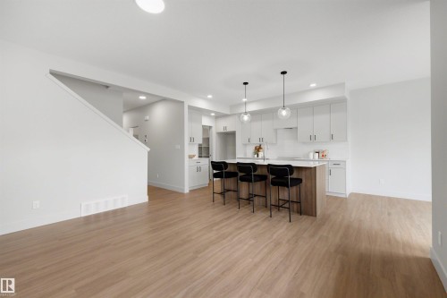 Open-concept living space featuring wood-finish flooring, a contemporary kitchen island with seating, pendant lighting, white cabinetry, and a white subway tile backsplash - 24 Grayson Green, Stony Plain, AB 