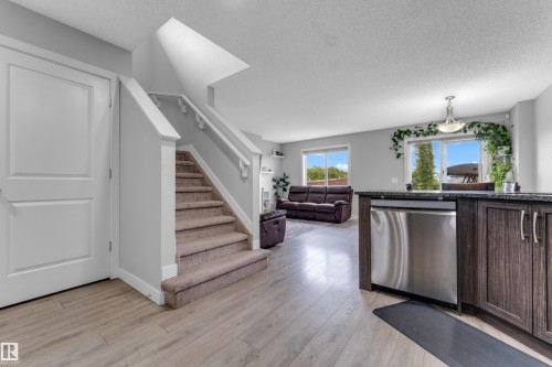 Open-concept living space featuring wood-finish flooring, a carpeted staircase with white railings, and a kitchen island with dark cabinetry and a stainless steel dishwasher - 1750 27 Street, Edmonton, AB 