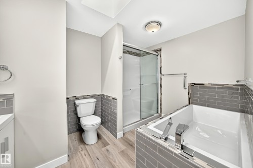 Bathroom featuring a glass-enclosed shower, a built-in tub with modern fixtures, wood-finish flooring, and gray subway tile wainscoting - 18636 71 Avenue, Edmonton, AB 