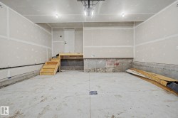 Spacious garage featuring a concrete floor, unpainted drywall, two ceiling-mounted light fixtures, and a wooden stairwell with a landing and white door - 