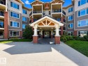 Contemporary multi-story building featuring brick and siding accents, multiple balconies with black railings, a prominent covered entrance with columns, and surrounding green landscaping - 117 3715 Whitelaw Lane, Edmonton, AB 