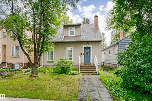 Traditional two-story residence with a stucco exterior, featuring a prominent blue entry door, white-trimmed windows, and a shingle roof with a dormer - 11105 81 Avenue, Edmonton, AB 