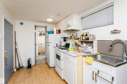 Compact kitchen featuring wood-finish flooring, white cabinetry, a built-in stainless steel sink with a high-arch faucet, and a window with horizontal blinds - 11105 81 Avenue, Edmonton, AB 