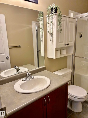 Bathroom featuring a vanity with a dark wood-finish cabinet and integrated sink, a large mirror, white wall-mounted cabinetry, a toilet, and a stand-up shower with a glass enclosure - 184 1804 70 Street, Edmonton, AB 