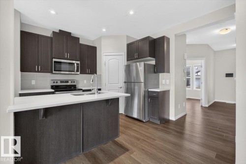 Modern kitchen featuring dark wood-finish cabinetry, stainless steel appliances, a center island with a sink, light-toned countertops, and wood-finish flooring - 2144 51 Street, Edmonton, AB 