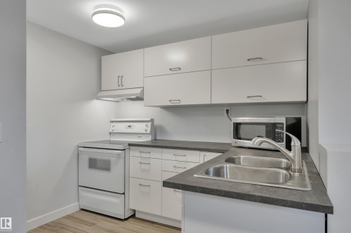 Kitchen featuring light-toned cabinetry with brushed nickel hardware, dark gray countertops, a stainless steel double-basin sink with a gooseneck faucet, a white electric range, and wood-finish flooring - 9835 74 Street, Edmonton, AB 