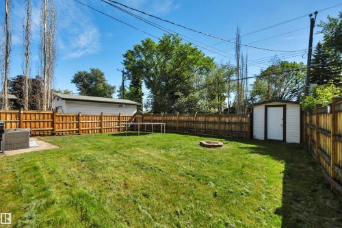 Expansive backyard featuring a manicured lawn, full wood fencing, a dedicated fire pit area, a white storage shed, and a concrete patio pad - 9835 74 Street, Edmonton, AB 