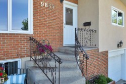 Front entrance featuring concrete steps with decorative wrought iron railings, brick facade, stucco siding, and a white entry door with a privacy screen insert - 