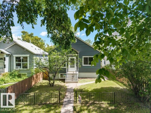 Siding exterior with a white entry door and concrete steps, featuring a wrought iron fence and mature landscaping - 11241 88 Street, Edmonton, AB 