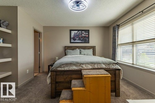 Bedroom featuring neutral wall paint, carpeted flooring, a large window with blinds, recessed floating shelves, and a ceiling-mounted light fixture with a fan - 17123 49 Street, Edmonton, AB 