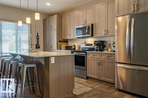 Modern kitchen featuring a breakfast bar island with seating, stainless steel appliances, wood-finish flooring, light-toned cabinetry, and pendant lighting - 17123 49 Street, Edmonton, AB 