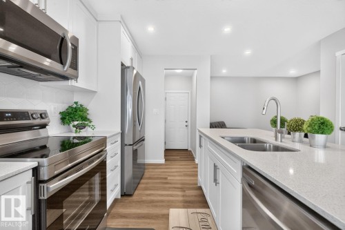 Modern kitchen featuring white shaker cabinetry, stainless steel appliances, a white geometric tile backsplash, wood-finish flooring, and a kitchen island with an undermount double sink - 1022 Hearthstone Boulevard, Sherwood Park, AB 