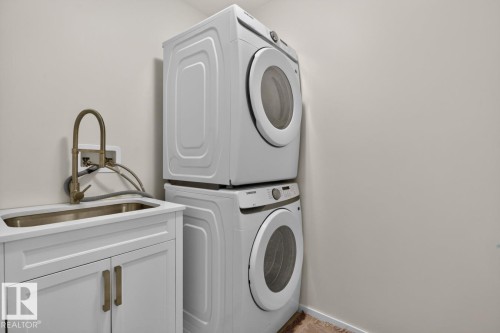 Dedicated laundry area featuring a stacked washer and dryer unit, a utility sink with a brushed bronze faucet, and white base cabinetry with matching hardware - 1022 Hearthstone Boulevard, Sherwood Park, AB 