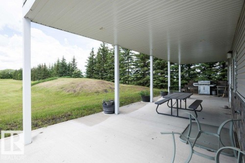 Covered concrete patio featuring white support columns, a textured ceiling, exterior siding with stone veneer accents, and a large grassy area with mature trees - 58505 Rng Rd 260, Rural Westlock County, AB 