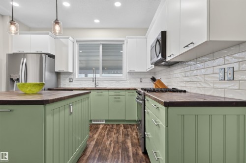 Kitchen featuring wood-finish flooring, two-tone cabinetry with white upper and green lower cabinets, wood countertops, and a white subway tile backsplash - 6928 22 Avenue, Edmonton, AB 