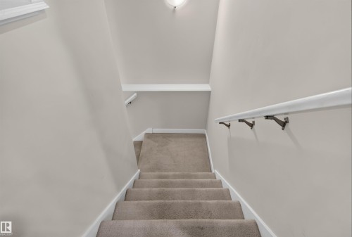 Carpeted staircase featuring a white handrail with dark bronze brackets, light neutral wall paint, white baseboards, and a recessed ceiling light fixture - 6928 22 Avenue, Edmonton, AB 