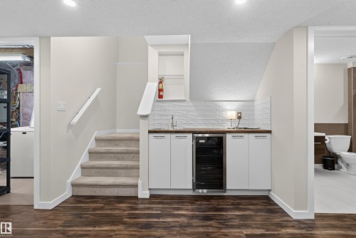 Wood-finish flooring anchors a space featuring a built-in wet bar with a compact beverage refrigerator, white cabinetry, and a textured white subway tile backsplash - 6928 22 Avenue, Edmonton, AB 