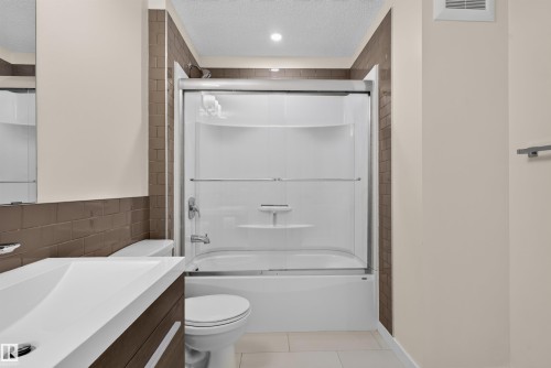 Bathroom featuring a shower-tub combination with clear sliding doors, a white integrated sink, a dark wood-finish vanity, and brown subway tile accents - 6928 22 Avenue, Edmonton, AB 