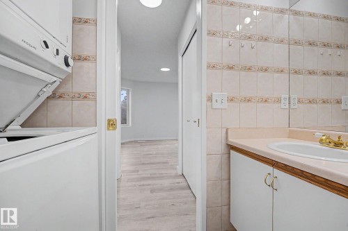 Integrated laundry appliances featuring a stackable washer and dryer, complemented by a vanity with a white basin and gold-tone faucet, set against a tiled wall with decorative accents and a full-width mirrored backsplash - 3704 39 Avenue, Edmonton, AB 