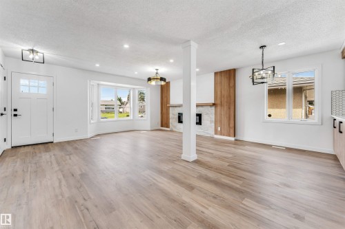 Spacious room featuring light wood-finish flooring, a contemporary fireplace with a stone surround and wood paneling, and an entry door with glass inserts - 6404 88 Avenue, Edmonton, AB 