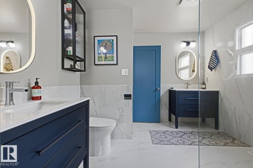 Dual vanity bathroom featuring an enclosed shower, white marble-look wall and floor tiling, and dark blue cabinetry - 3504 102 Avenue, Edmonton, AB 