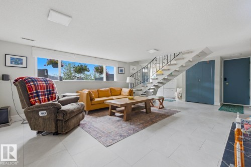 Spacious living area featuring light-toned tile flooring, a prominent staircase with wrought iron railing, a wide window span, built-in storage under the staircase, and a solid-panel entry door - 3504 102 Avenue, Edmonton, AB 