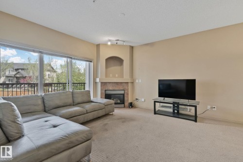 Living area with corner gas fireplace featuring a stone surround and mantel, sliding glass doors opening to a balcony, and neutral carpeting - 209 5280 Terwillegar Boulevard, Edmonton, AB - Indoor Photo Showing Living Room With Fireplace