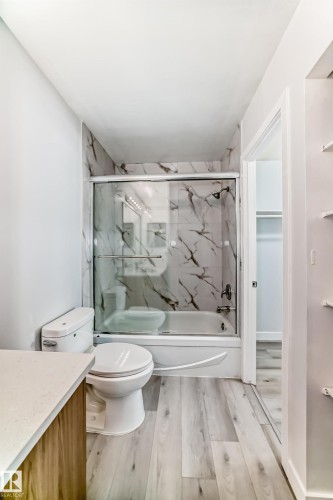 Bathroom featuring a tub/shower combination with sliding glass doors and marble-patterned tile surround, a toilet, wood-finish flooring, and a vanity with a light-colored countertop - 23 1503 Mill Woods Road E, Edmonton, AB 