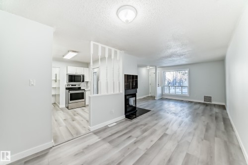 Bright living area featuring wood-finish flooring, a modern fireplace with a dark surround, a large picture window, and neutral wall tones - 23 1503 Mill Woods Road E, Edmonton, AB 