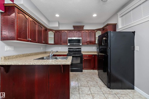 Kitchen featuring dark wood cabinetry, light-toned countertops, a double basin stainless steel sink, and a full suite of black appliances, including a microwave and refrigerator - 4831 154 Avenue, Edmonton, AB 