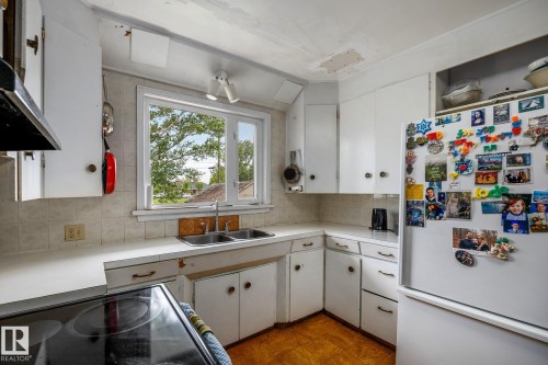 Kitchen featuring white cabinetry, a double basin stainless steel sink, a window overlooking exterior greenery, and tile flooring - 11022 110 Avenue, Edmonton, AB 