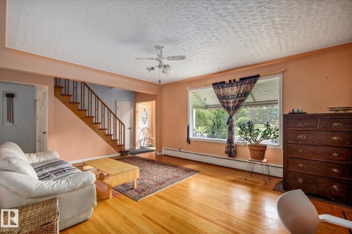 Bright living space featuring wood-finish flooring, a prominent staircase with wood railing, a ceiling fan, and a large front-facing window - 11022 110 Avenue, Edmonton, AB 