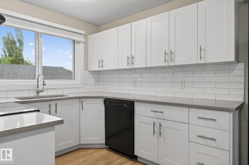 Modern kitchen featuring white shaker-style cabinetry, a stainless steel sink with a gooseneck faucet, light grey countertops, a black built-in dishwasher, and wood-finish flooring - 15112 80 Street, Edmonton, AB 