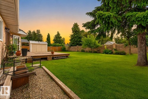 Expansive backyard featuring a manicured lawn, a large wood deck, mature evergreen trees, a wood privacy fence, and a brick wall - 9 Emerald Terrace, St. Albert, AB 