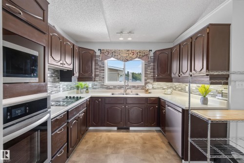 U-shaped kitchen featuring dark wood cabinetry, white countertops, a mosaic tile backsplash, and stainless steel appliances including a built-in microwave and oven - 10935 152 Street, Edmonton, AB 