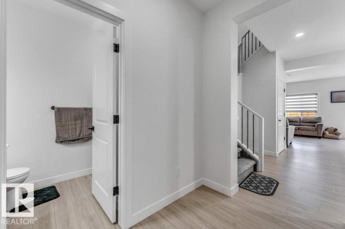 Bright hallway with wood-finish flooring, a white interior door with black hardware, and a staircase featuring carpeted treads and black metal balusters - 423 28 Street, Edmonton, AB 
