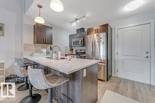 Modern kitchen featuring dark wood-finish cabinetry, stainless steel appliances, a white countertop island with a built-in sink, and wood-finish flooring - 401 667 Watt Boulevard, Edmonton, AB 