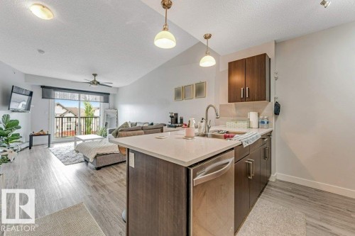 Open-concept living space featuring a modern kitchen island with an integrated stainless steel dishwasher, wood-finish flooring, a sliding glass door to an exterior balcony, and a ceiling fan - 401 667 Watt Boulevard, Edmonton, AB 